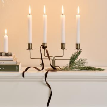 A brass four arm advent candelabra holding five lit white taper candles, styled on a white mantel with a dark velvet ribbon, pine sprigs, and stacked books against a warm beige wall.